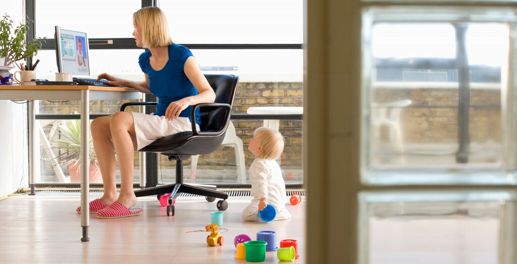 Young woman working on a computer in her home office with her baby girl on the floor playing with toys and looking up at her mother