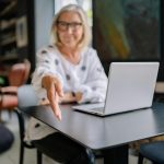 middle-aged woman at a table with a laptop, extending her hand for a handshake